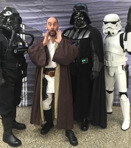 David Engel is dressed as a jedi in brown robes and a white shirt. He is surrounded by two storm troopers and Darth Vader is standing behind him.