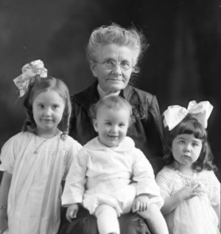 Black and white photograph of an old woman, two young girls with white bows in their hair and a baby boy.