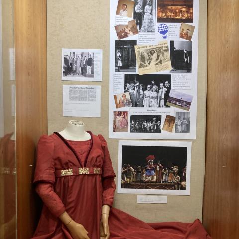 Mannequin wearing empire waist red dress in exhibit of Lake George Opera Festival. Photos of various operas appear next to the costume.