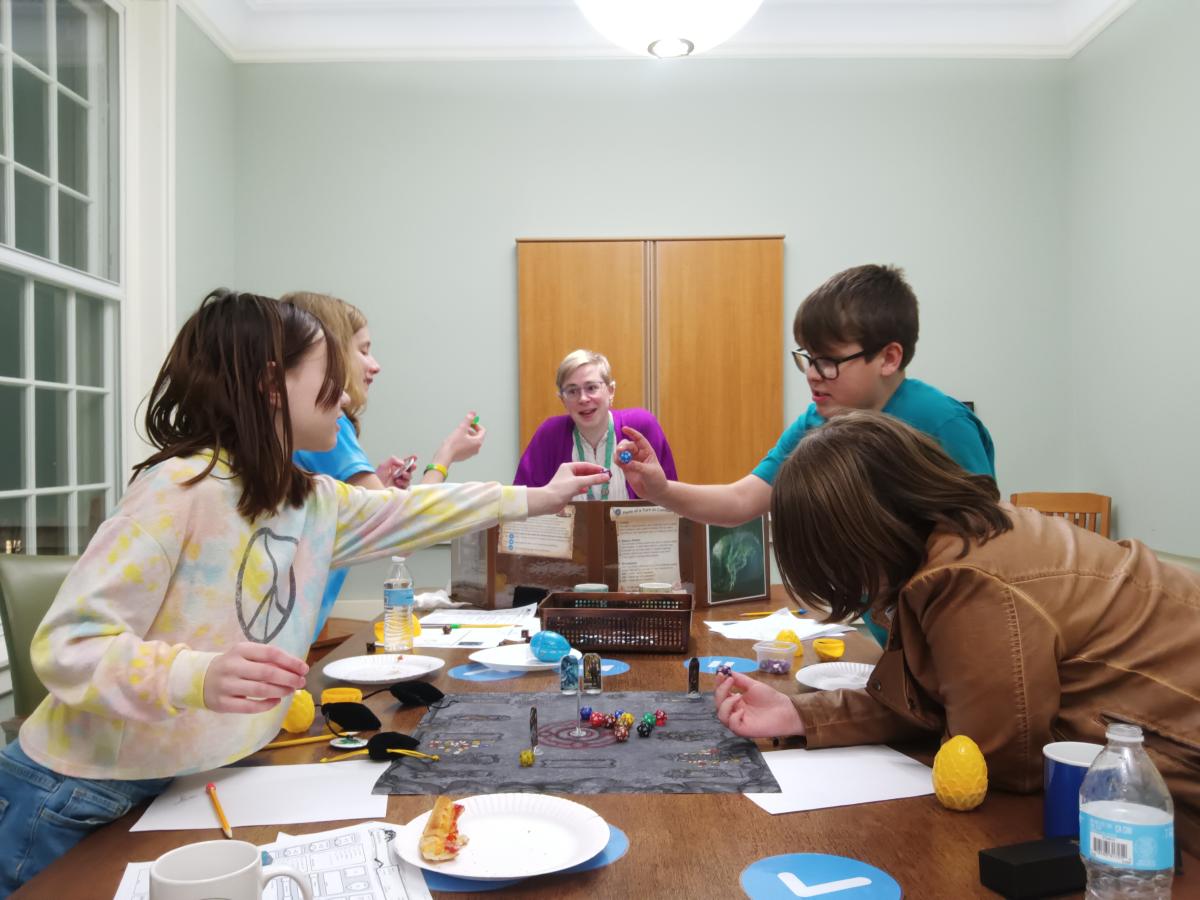 Four tweens and one adult standing around a long rectangular table. The table has maps, dice, and food set on it. The tweens are all holding dice.