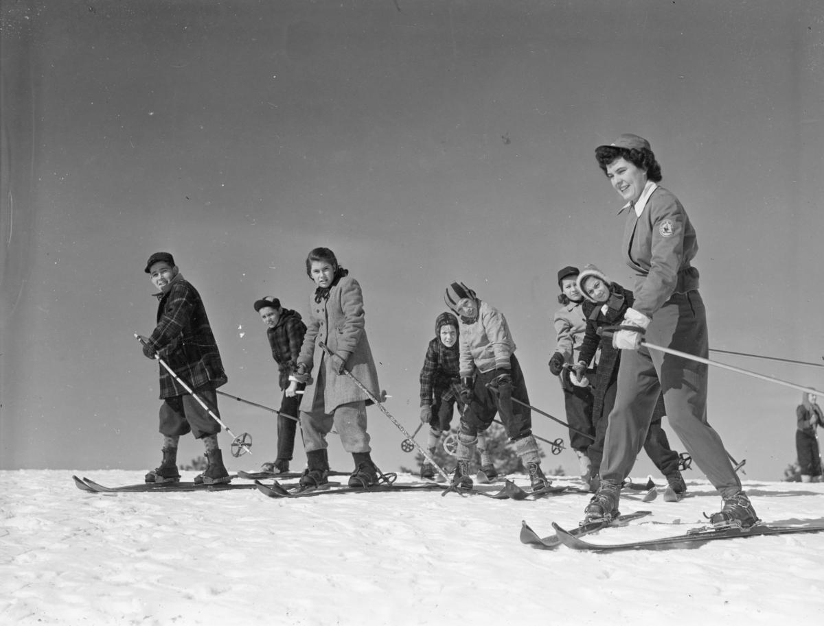 Black and white photograph of a woman on skis teaching skiing safety a group of kids on skis.